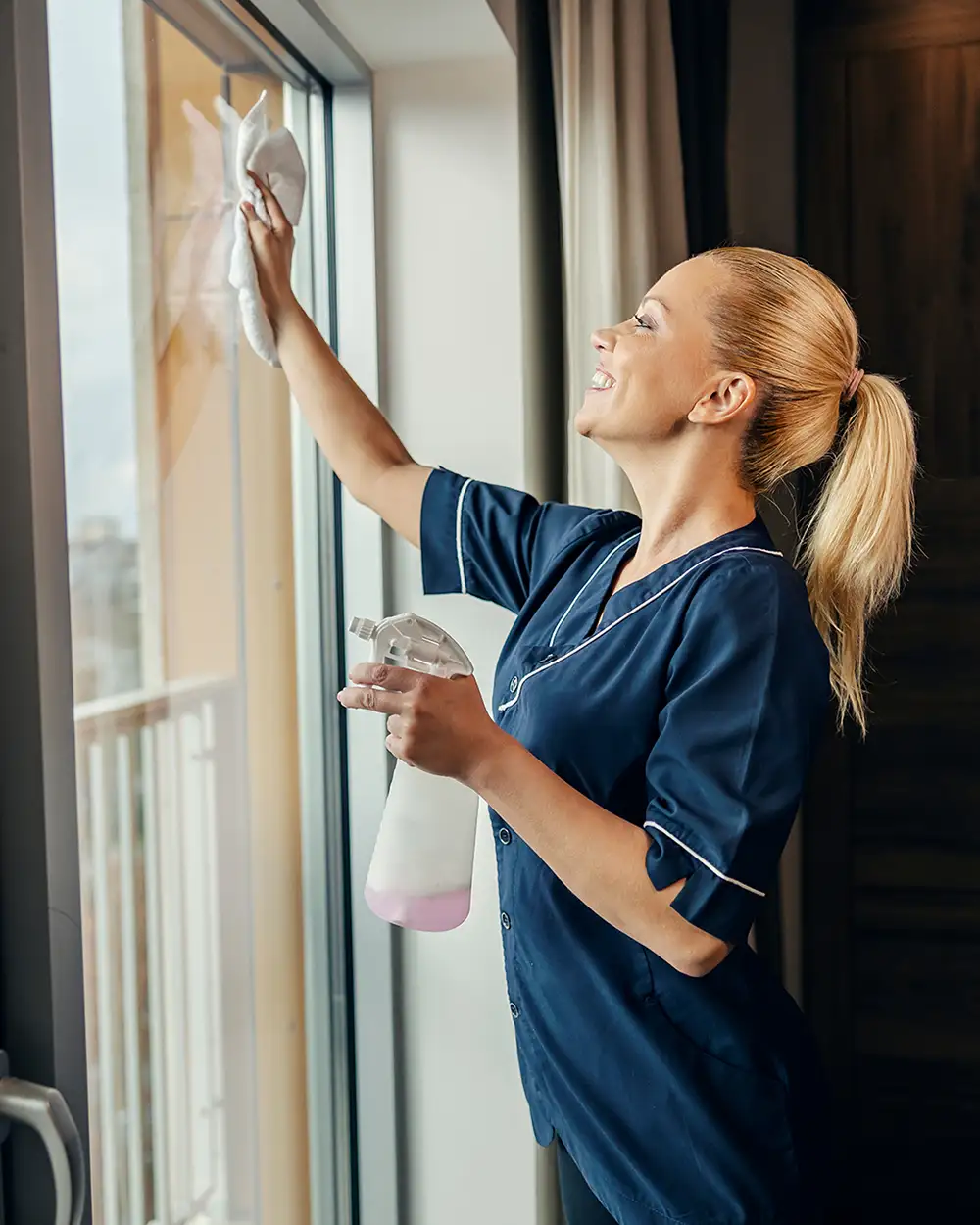 Ein glückliches Zimmermädchen putzt die Fenster eines Hotelzimmers mit Reinigungsmittel und Lappen.