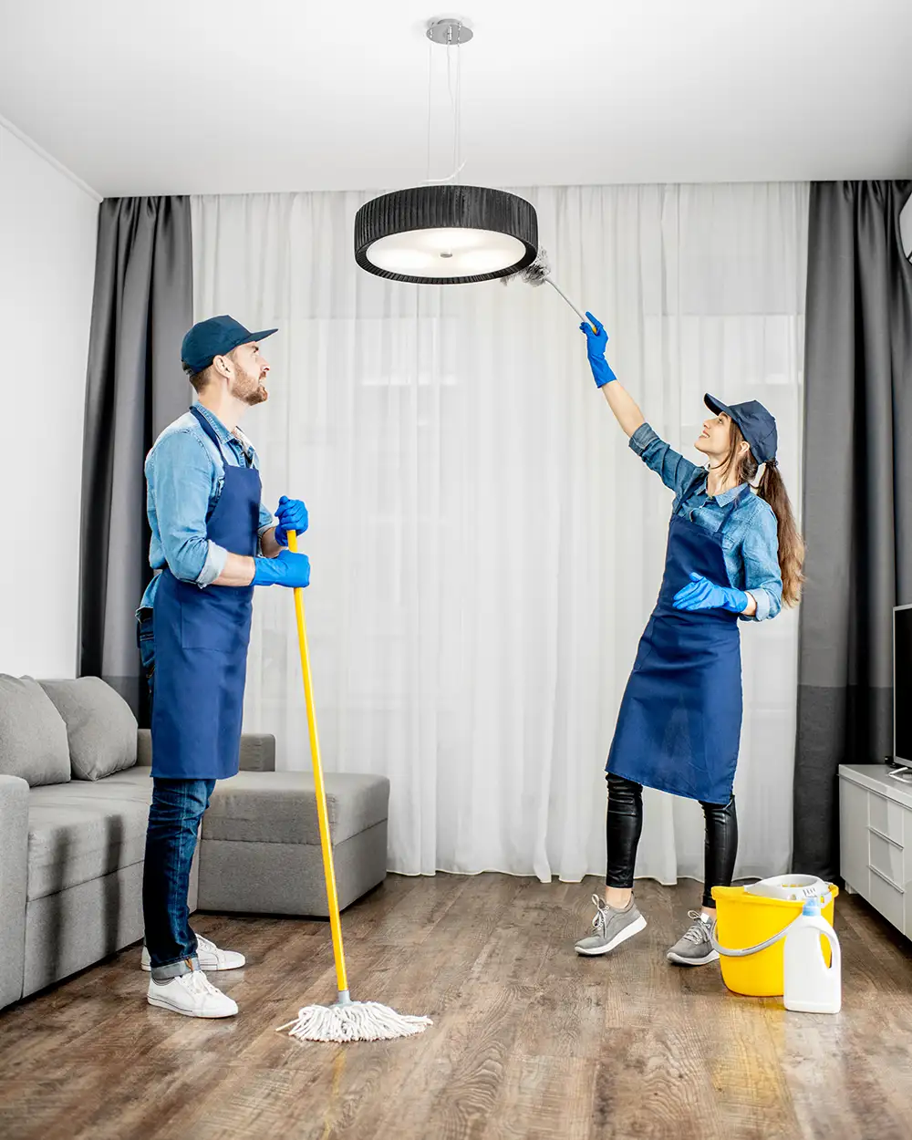 Professionelle Reinigungskräfte in blauer Uniform waschen den Boden und wischen den Staub von der Lampe im Wohnzimmer der Wohnung.